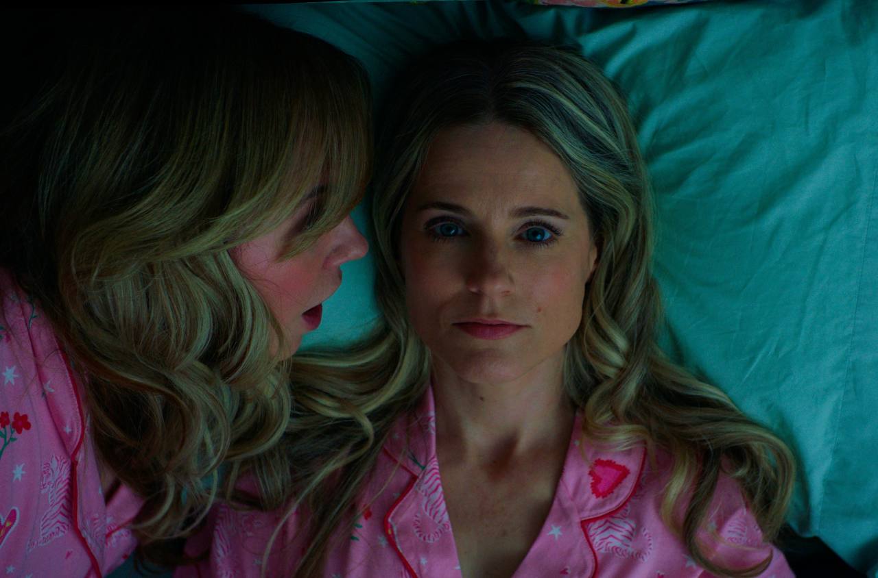 Two women in matching pink pajamas lie together on teal bedding as one whispers to the other.