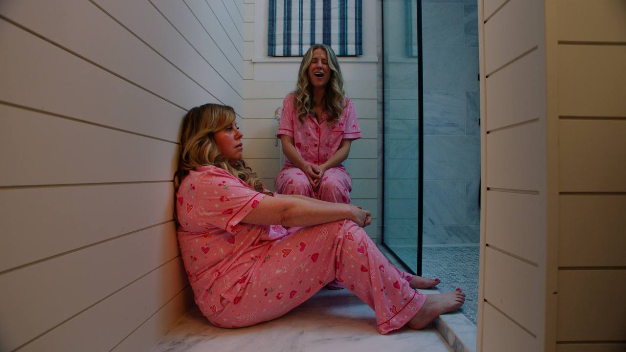 Two women in pink pajamas sit on a narrow bathroom floor as one cries out.