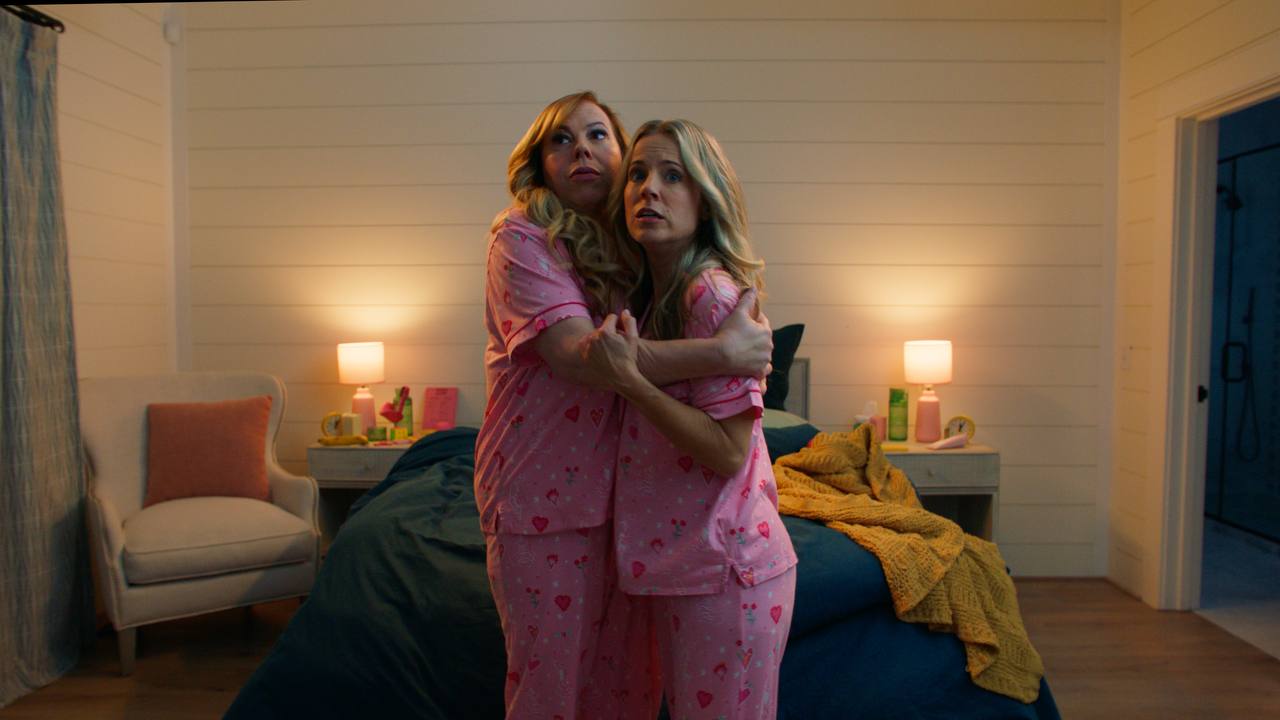 Two frightened women in matching pink pajamas embrace in a bedroom.
